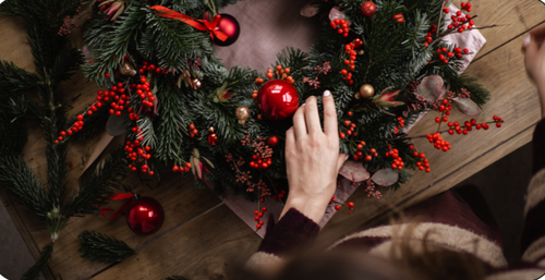 Decorative wreath with red ornaments on a wooden surface, with a hand adjusting the wreath. Floral Workshop