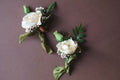 Two floral boutonnieres with white roses and green leaves on a brown background. Designed by The Dreamt Garden in Madison, AL for small weddings.