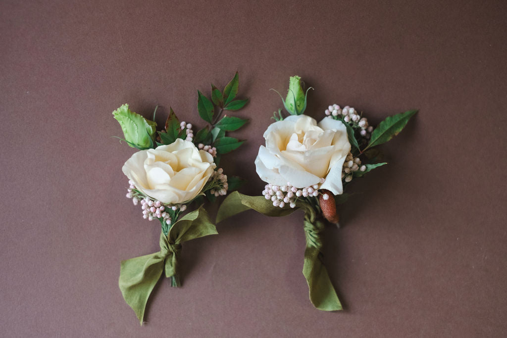 Two floral boutonnieres with white roses and green leaves on a brown background.  For Prom, Homecoming, and Formals. Whimsical flowers by modern retail flower shop in Madison AL.