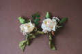 Two floral boutonnieres with white roses and green leaves on a brown background.  For Prom, Homecoming, and Formals. Whimsical flowers by modern retail flower shop in Madison AL.