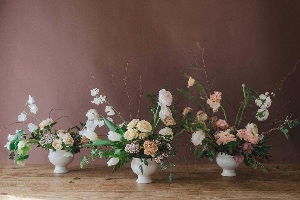Three floral arrangements with roses lisianthus tulips in white vases on a wooden surface with a brown background.  From The Dreamt Garden, a retail flower and garden store in charming historic downtown Madison AL.