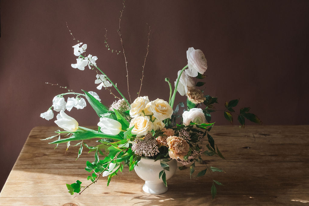 Floral arrangement with Roses carnations sweet peas vines and nandina in a white vase on a wooden table.  Cottage and garden inspired flowers from The Dreamt Garden.