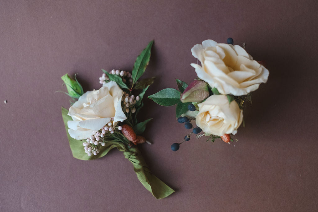 Floral boutonniere and corsage with white roses and green leaves on a brown background. Designed by The Dreamt Garden in Madison, AL for small weddings.