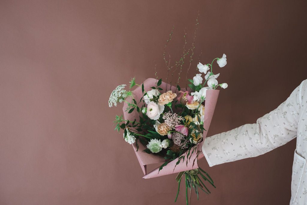 A hand holding a bouquet of mixed wildflowers wrapped in pink paper, set against a solid dusty rose background.