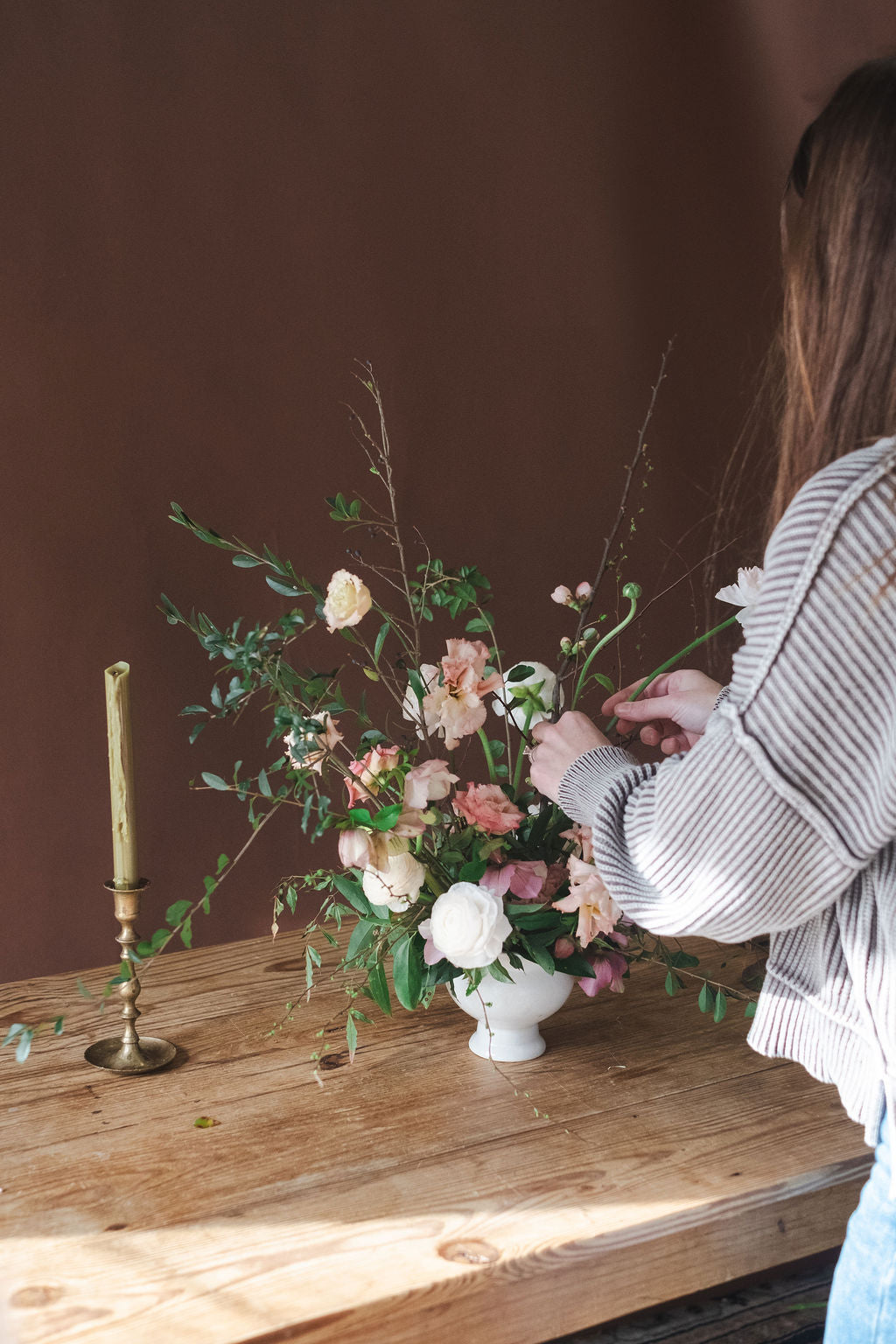 The Dreamt Garden florist in Madison Huntsville Alabama arranging a floral bouquet in a white vase on a rustic wooden table, with a brass candlestick holder beside it against a dark brown wall.  Garden style floral arrangement for corporate events, baby showers, bridal showers, rehearsal dinners, holiday flowers, thanksgiving flowers, Christmas flowers.