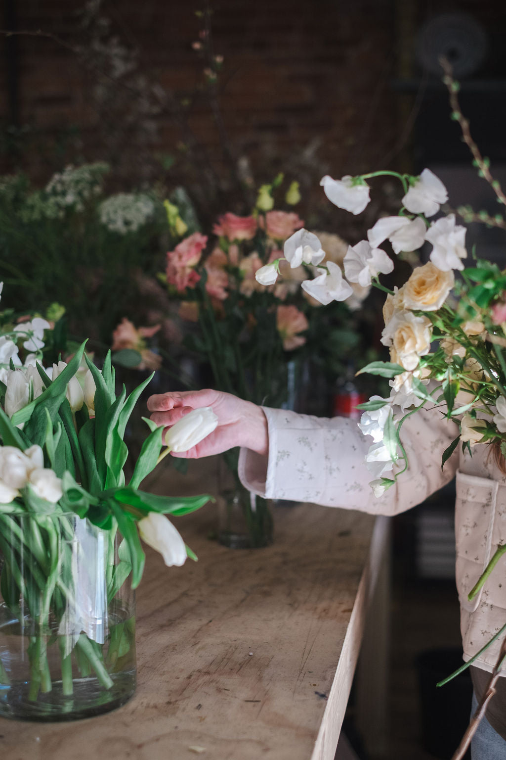 Madison AL florist arranging white tulips in a clear glass vase on a rustic wooden table, with other bouquets of white, pink, and yellow flowers in the background of a dimly lit flower shop.  Teaching flower arranging workshop and wreath workshop Madison AL Huntsville, Alabama.