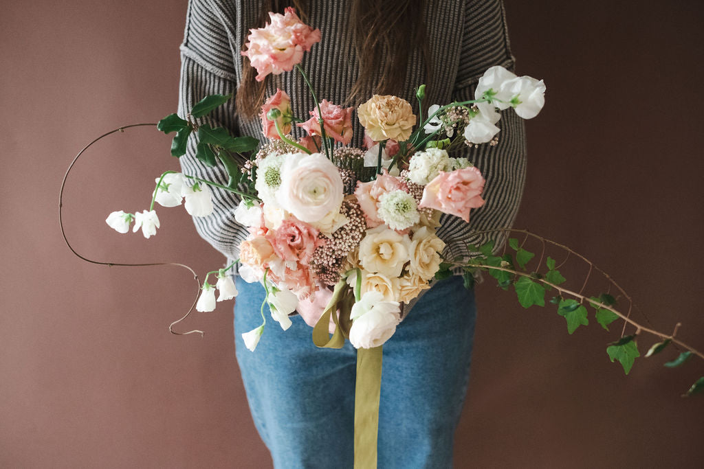 The Dreamt Garden Madison AL florist holding a large, elaborate floral bouquet with pastel pink, peach, cream, and white flowers, green foliage, and a green ribbon, set against a solid dark pinkish brown background.